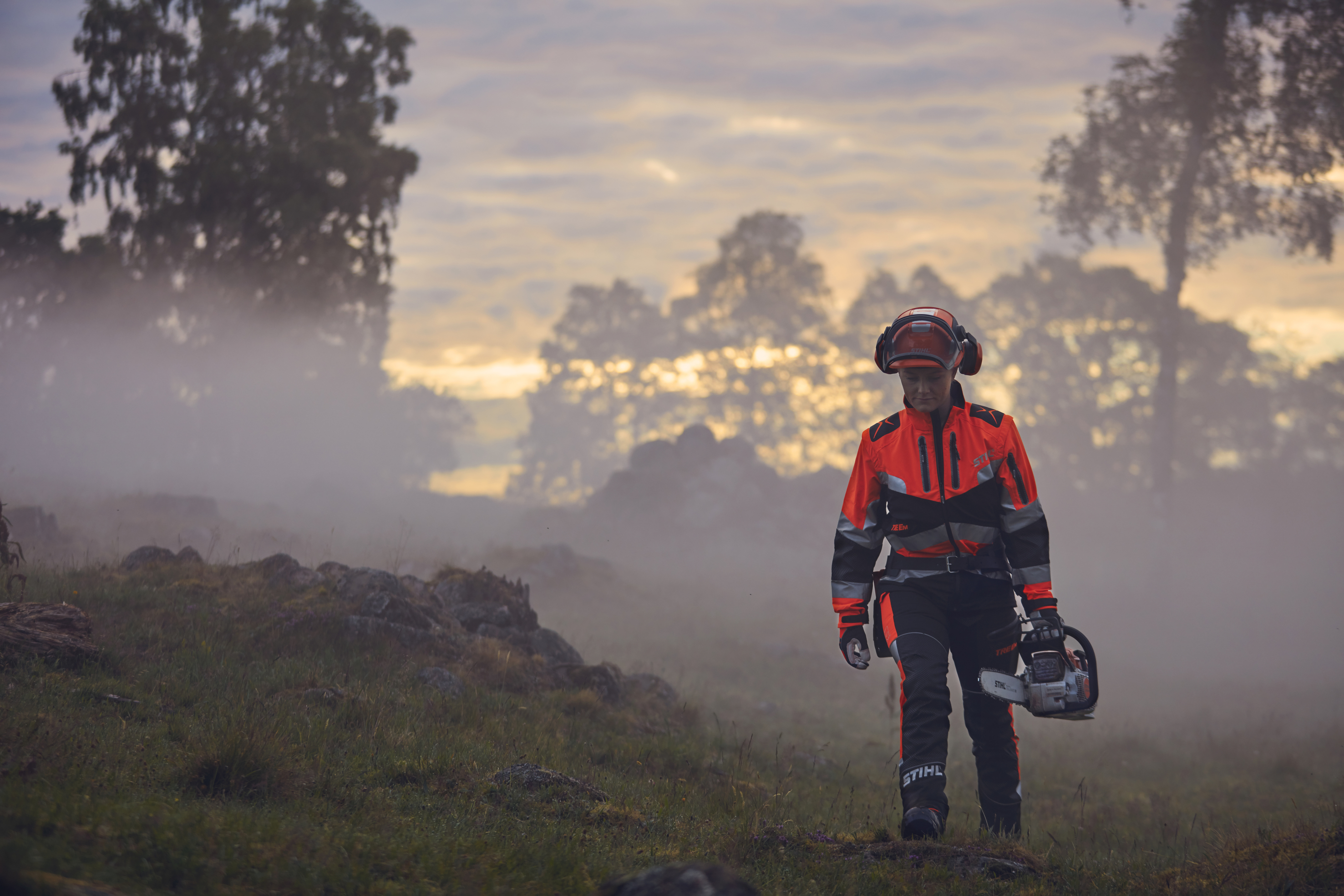 En kvinnelig skogsarbeider går kledd i STIHL arbeidsklær i soloppgang på en høyde
