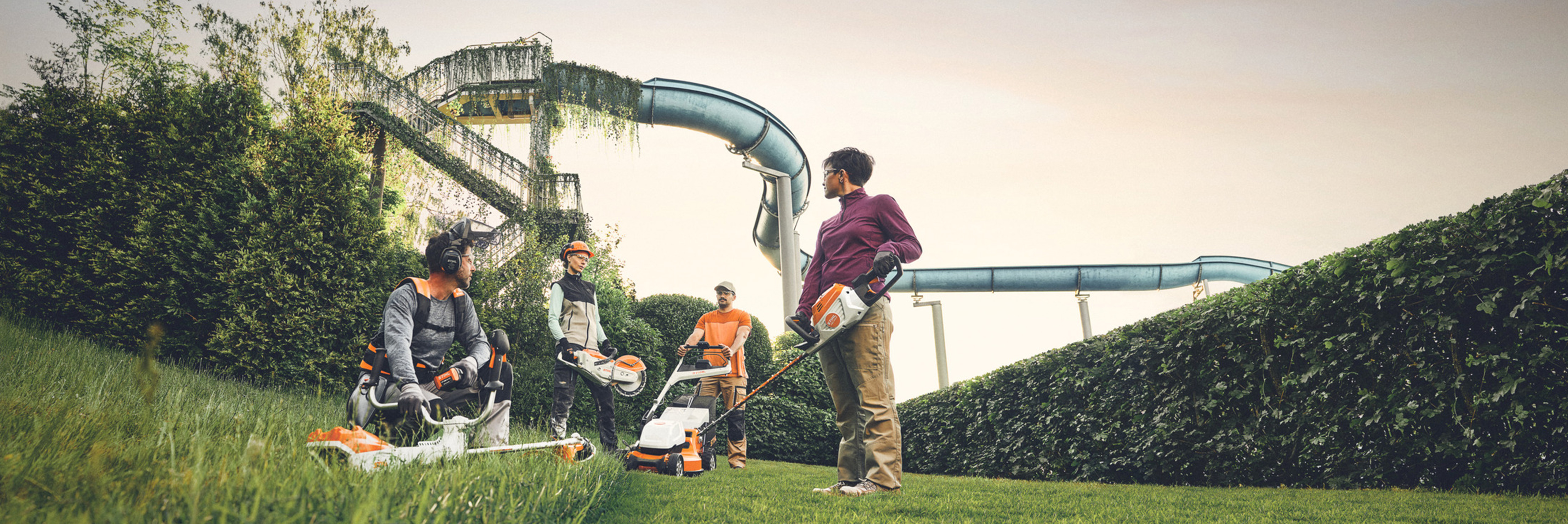 Een groep mensen werkt met STIHL tuingereedschap op een groen gebied met een grote haag en waterglijbanen op de achtergrond. Ze zijn uitgerust met bosmaaiers, grasmaaiers en beschermende uitrusting.