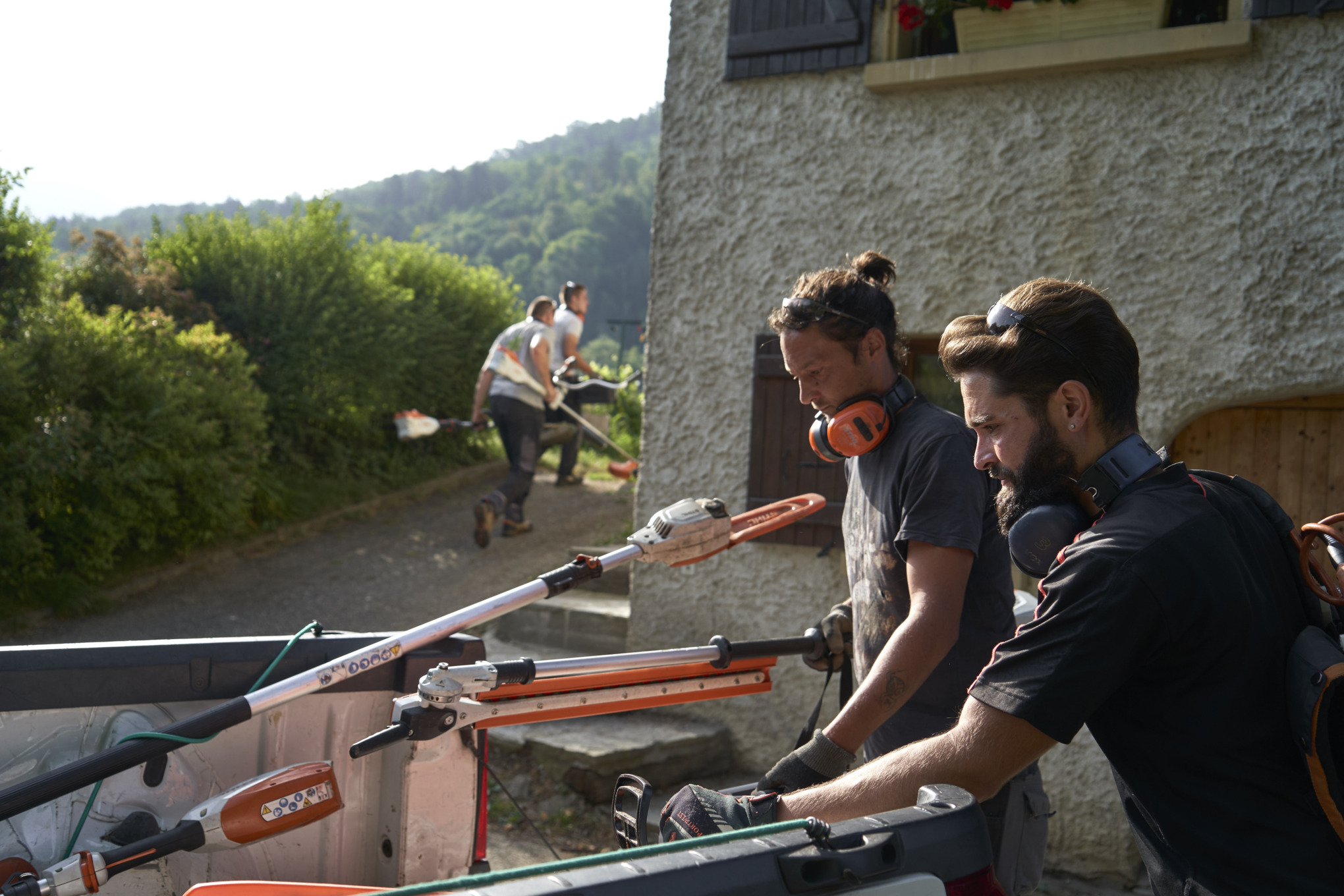 To anleggsgartnere laster ut STIHL-batteriverktøy fra en bil, med andre arbeidere i bakgrunnen 