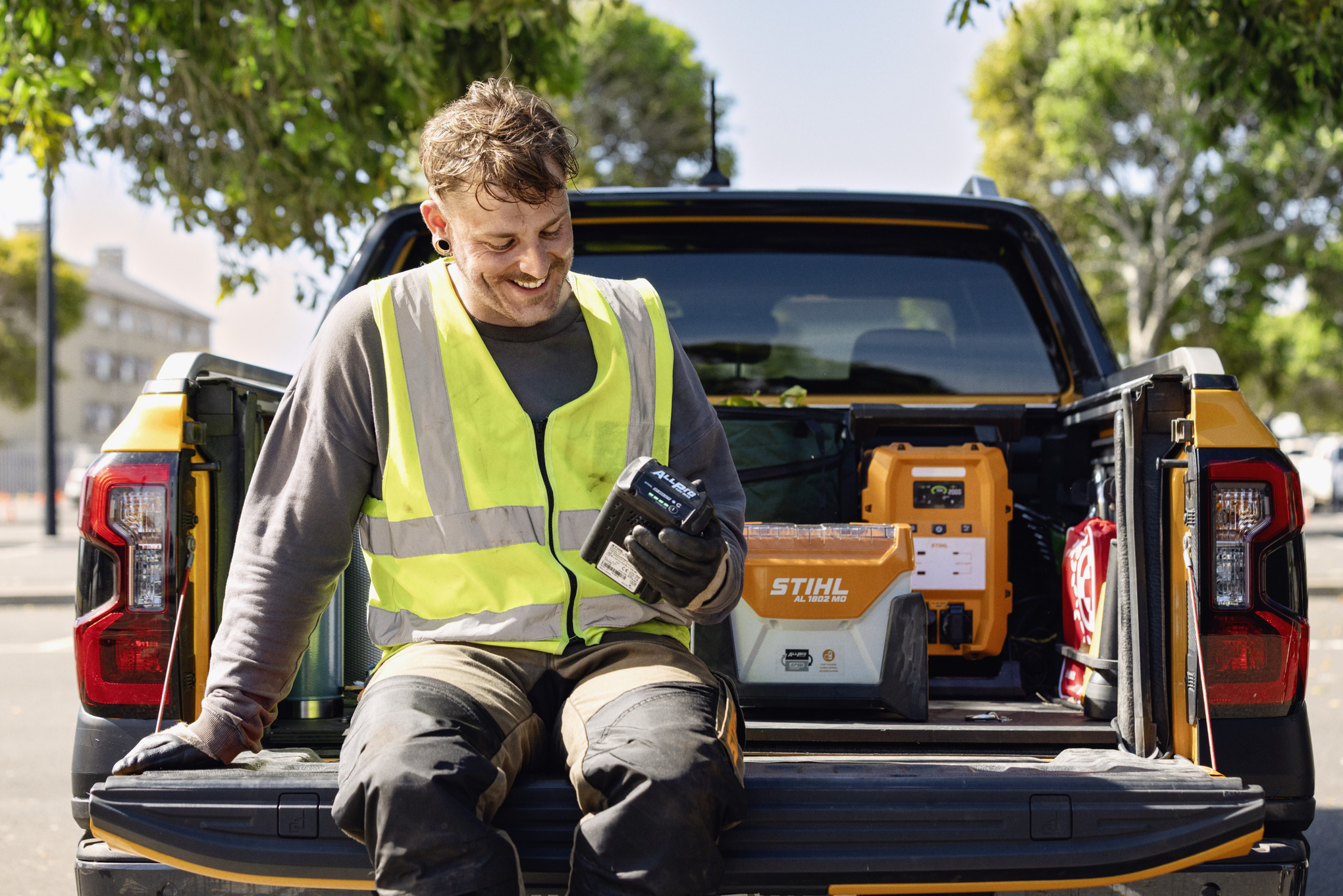 Mann som sitter på baklemmen til en pickup og sjekker ladenivået på sitt ALLPRO-batteri. I bakgrunnen er hurtigladeren STIHL AL 1802 MO og den mobile strømforsyningen PS 3000