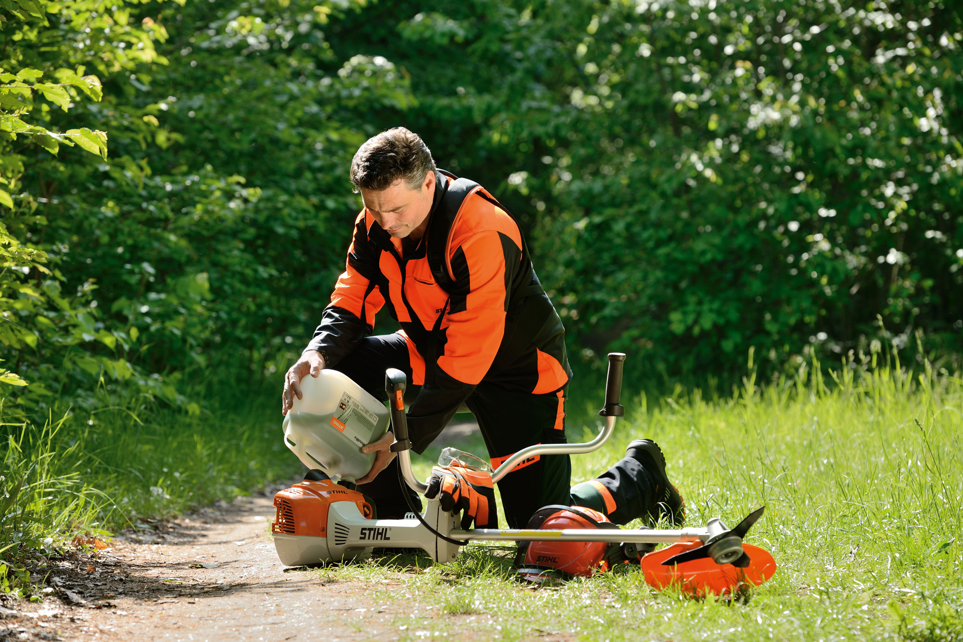 En mann står foran noen busker og fyller drivstofftanken til en STIHL gresstrimmer med en stor flaske