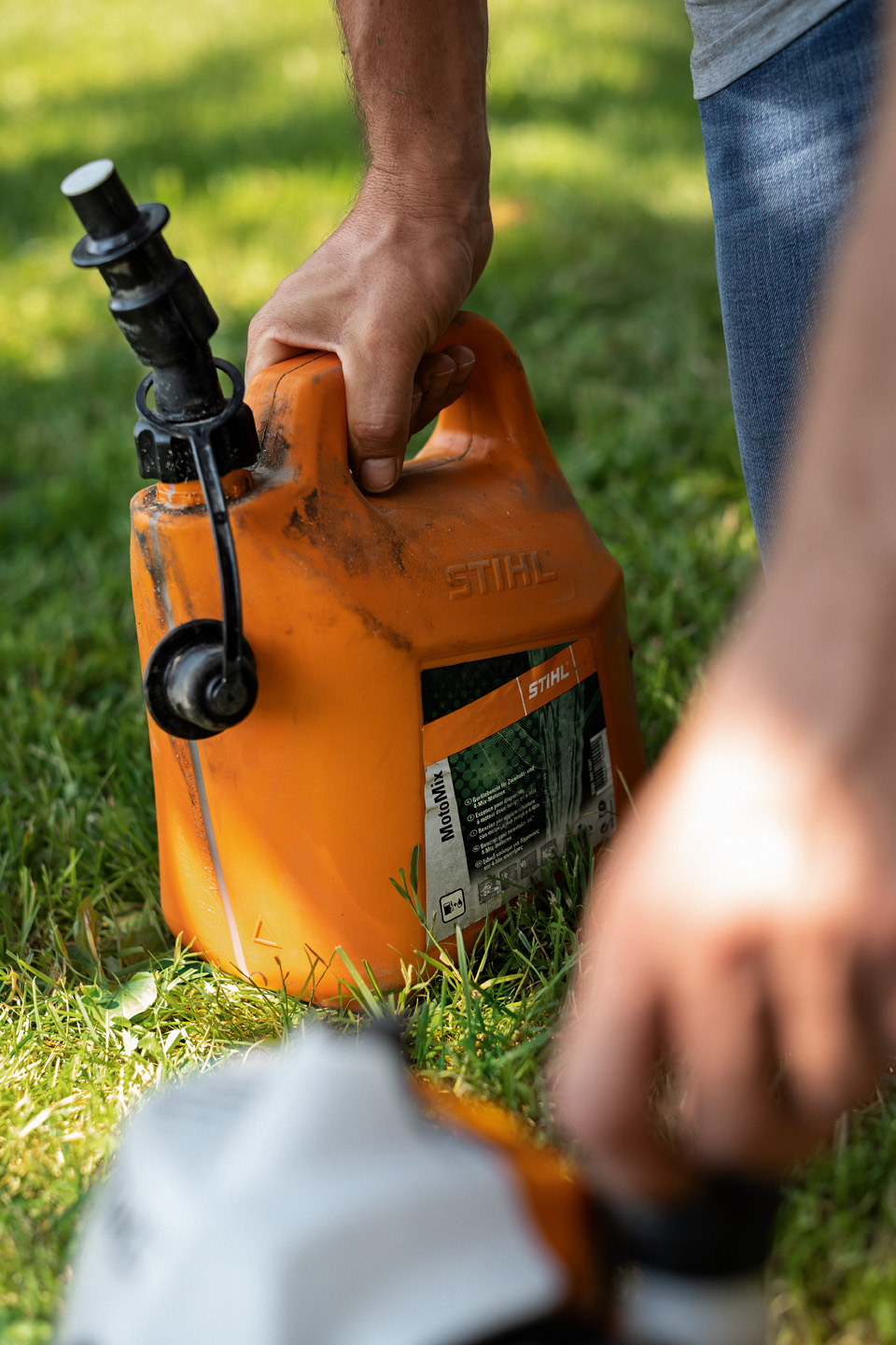 En skitten kanne med STIHL MotoMix drivstoffblanding på gress, med en hånd som åpner tanklokket på et verktøy i forgrunnen