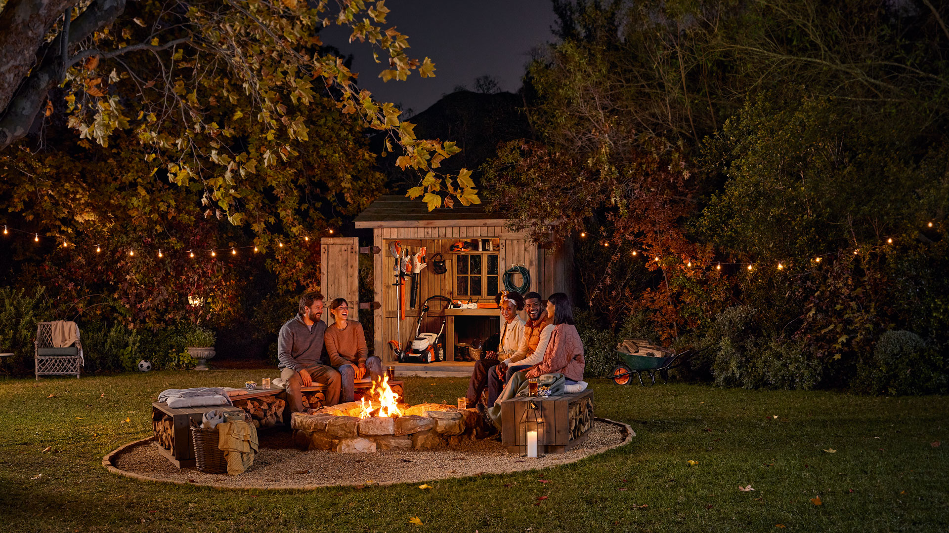 3 kvinnor och 2 män sitter samlad kring en öppen eldstad i trädgården bland STIHL-verktyg i skymning