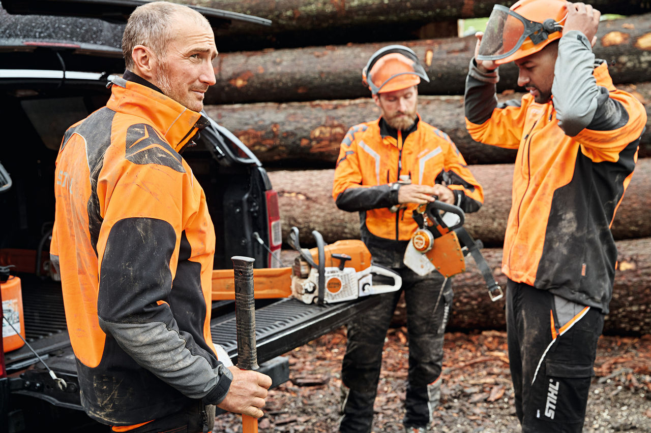 Tre menn i personlig verneutstyr fra STIHL holder i batteridrevne verktøy ved siden av et åpent bagasjerom foran en tømmerlunne