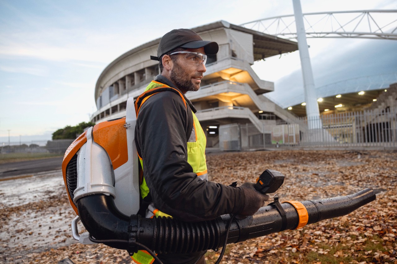 Sent på kvelden rydder en person løv og rusk fra et stadion ved hjelp av den batteridrevne løvblåseren ALLPRO STIHL BRA 600.